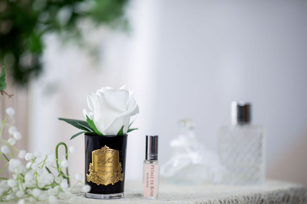 
                  
                    A white rosebud in a black glass vase with gold crest, beside baby's breath flowers on a dressing table, covered with a white lace cloth.  Sitting beside that is a rose de petale fragrance spray to scent the flowers.  These are displayed at Natashas Skin Spa beauty salon and florist on city road southbank in melbourne australia
                  
                