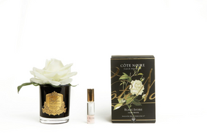 
                  
                    Cote Noire single rose, open and full, in ivory white with a black glass vase decorated with a gold crest, sits beside a bottle of rose petal perfume to spray on it's petals, and a pretty gift box in black, gold and white, as seen at Natashas Skin Spa Southbank
                  
                